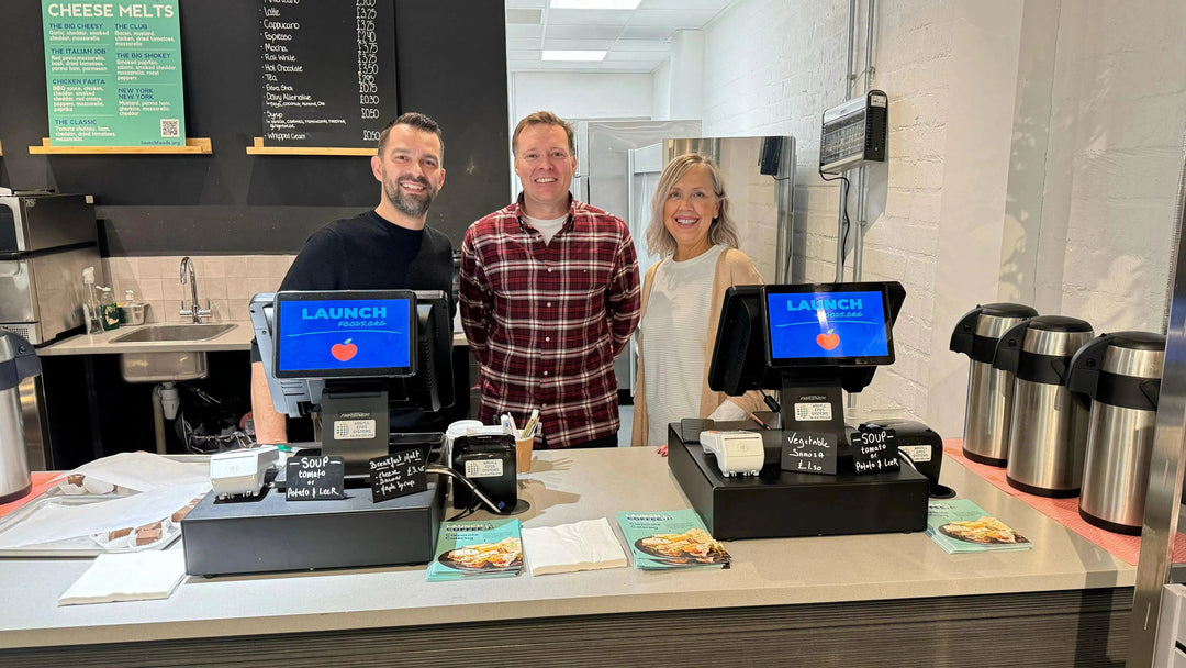 Three staff stand smiling behind the Launch Foods cafe counter with menu boards and tills visible.