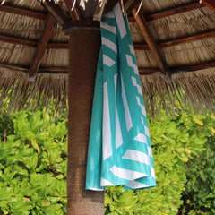 Turquoise and white geometric beach towel hanging on a tiki hut post with leafy greenery.