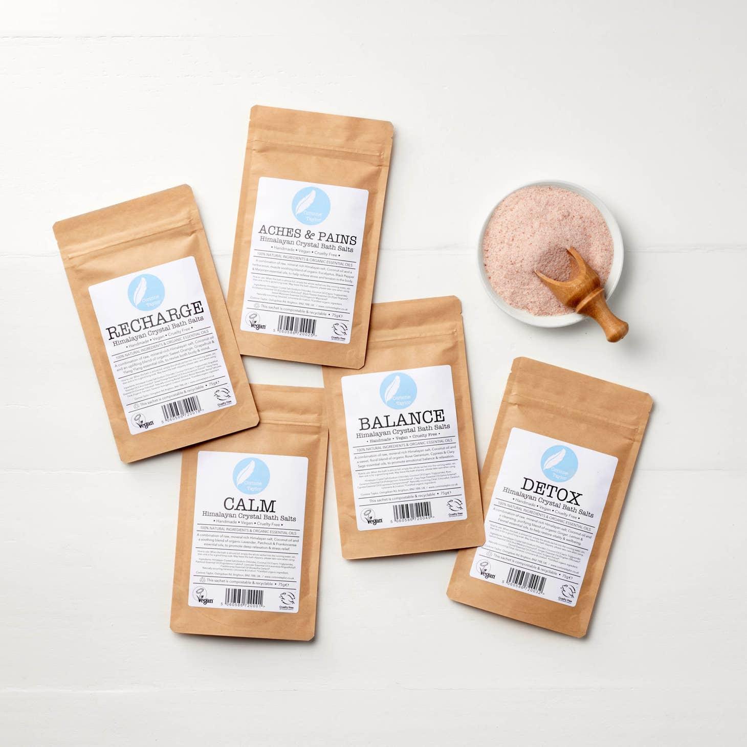 Five Himalayan Crystal Bath Salts pouches with wooden scoop and bowl of pink salt on white table.