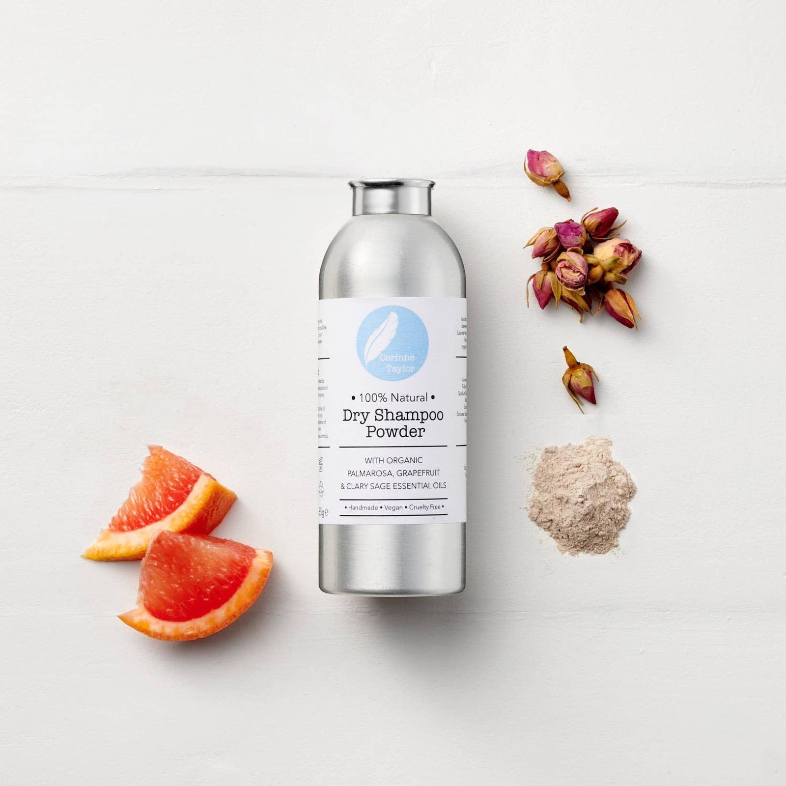 100% natural dry shampoo powder bottle with grapefruit slices, dried rosebuds, and powder on white background