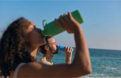 People drinking from reusable water bottles on a sunny beach with blue ocean background