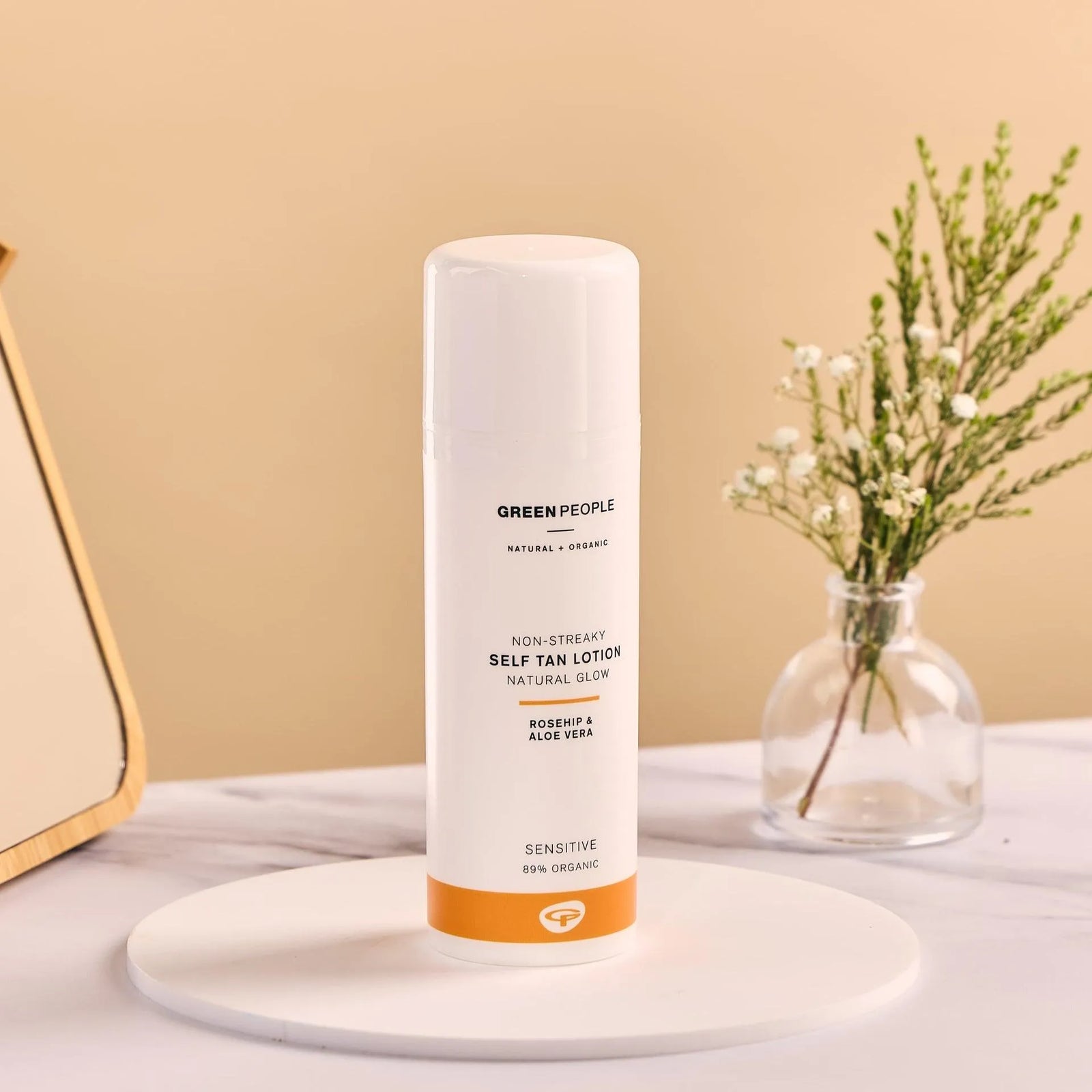 Green People self tan lotion bottle on white stand with small glass vase and flowers