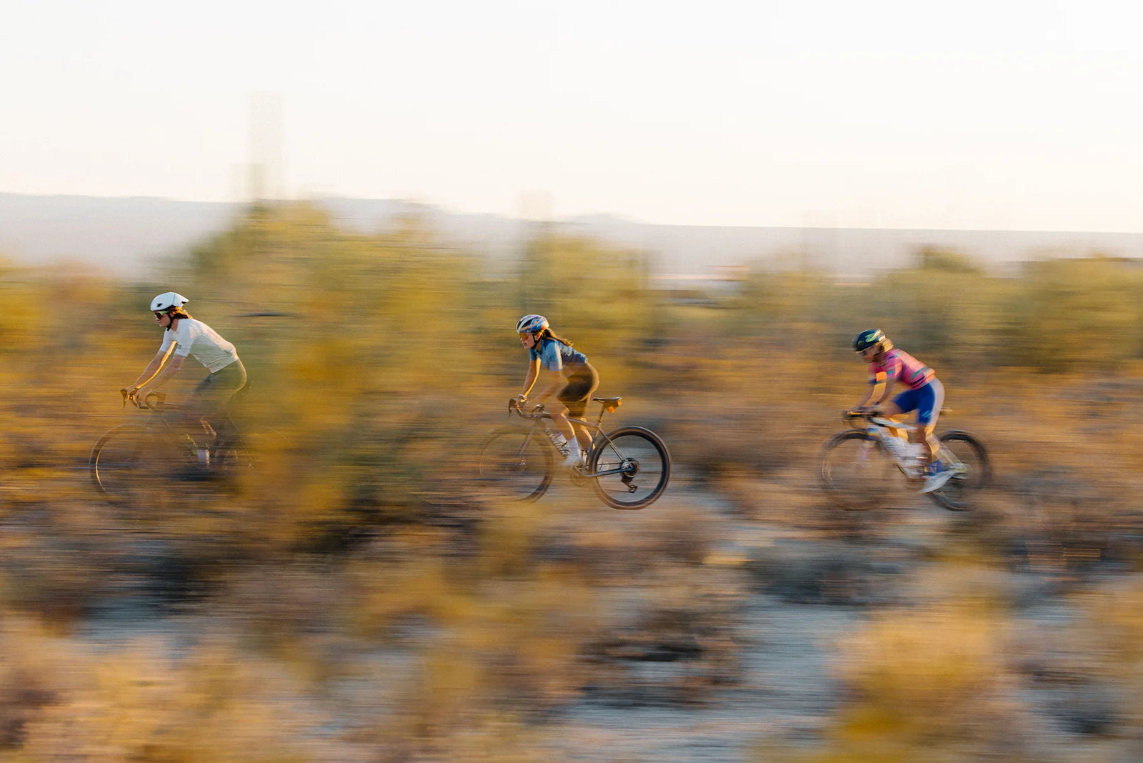 Three cyclists ride gravel bikes off-road in a desert landscape at sunset