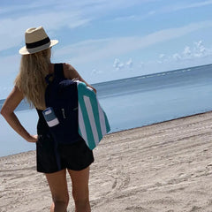 Woman with backpack and striped beach towel standing on sandy beach by the ocean