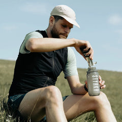Man outdoors mixing STYRKR energy drink powder into water bottle on grassy hill