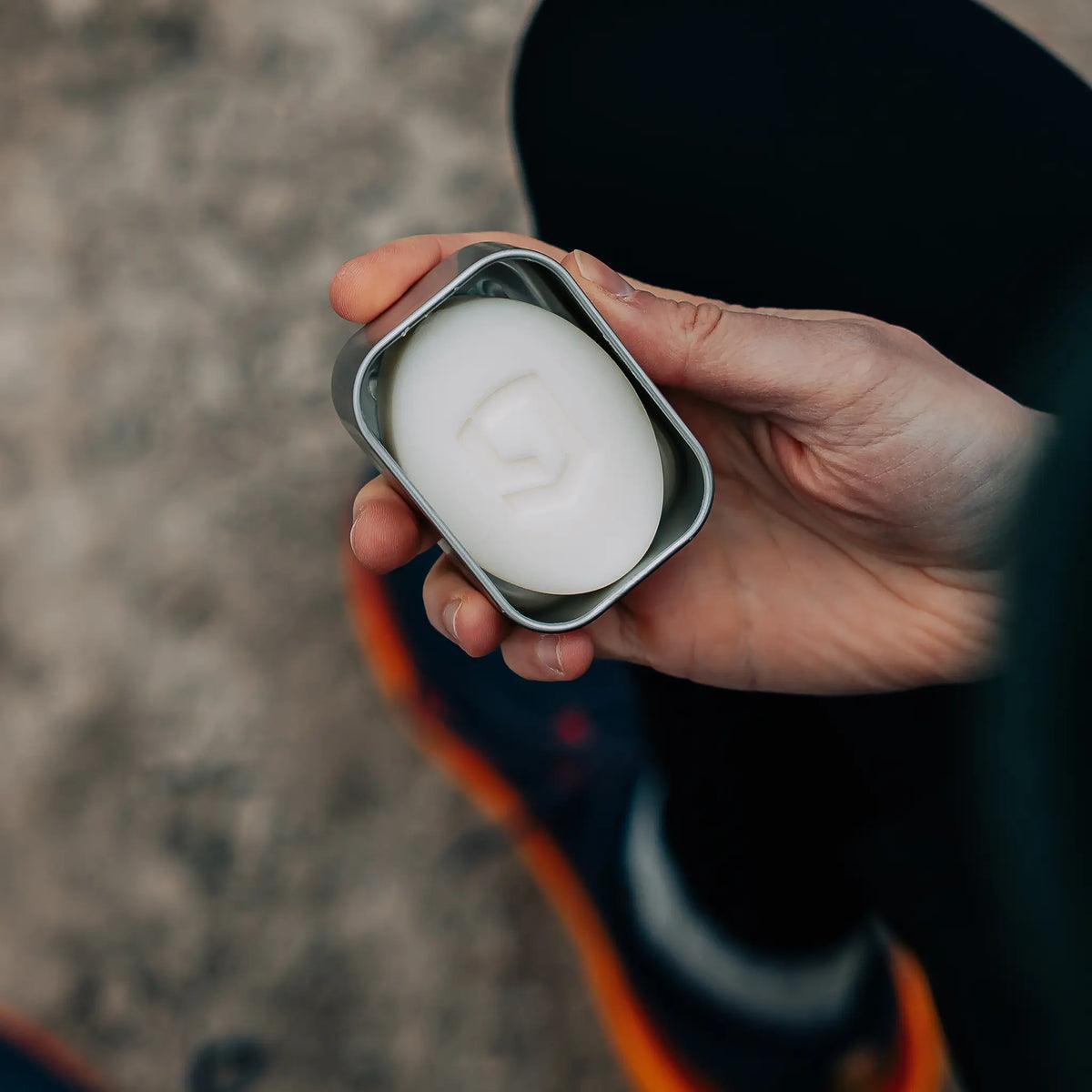 Hand holding white anti-chafe bar in metal tin, outdoor setting, running shoe visible