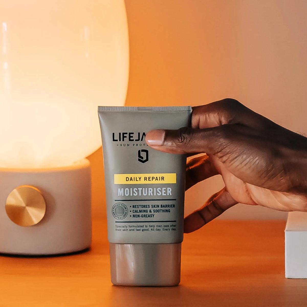 Hand holding LifeJacket Daily Repair Moisturiser tube on wooden surface by lamp