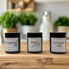 Trio of Ready Sweat Go soy candles—Coastal Cottage, Sage + Sea Salt, Balsam + Cedar—on wood table