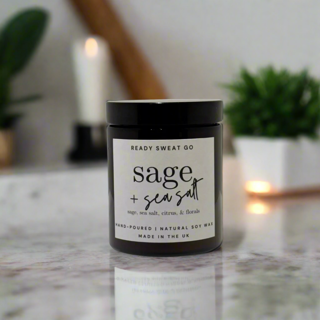 Sage and sea salt soy candle in black jar on marble surface, with blurred plants and candle in background