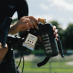 Cyclist holding STYRKR water bottle and protein rice bar outdoors on a bike