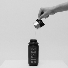 Hand pouring STYRKR SLT05 powder into black water bottle, minimalist studio setting