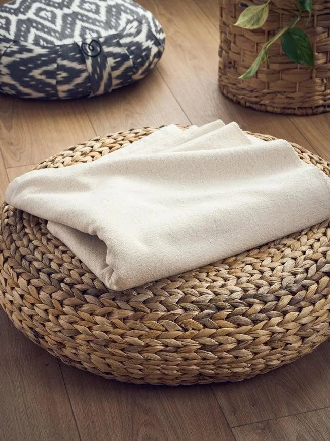 Folded beige blanket on round woven ottoman with cushion and wicker basket on wooden floor