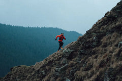 Trail runner in red jacket running uphill on a rocky mountain with forest background