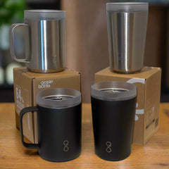 Stainless steel and black Ocean Bottle mugs and tumblers on cardboard boxes, wooden table.