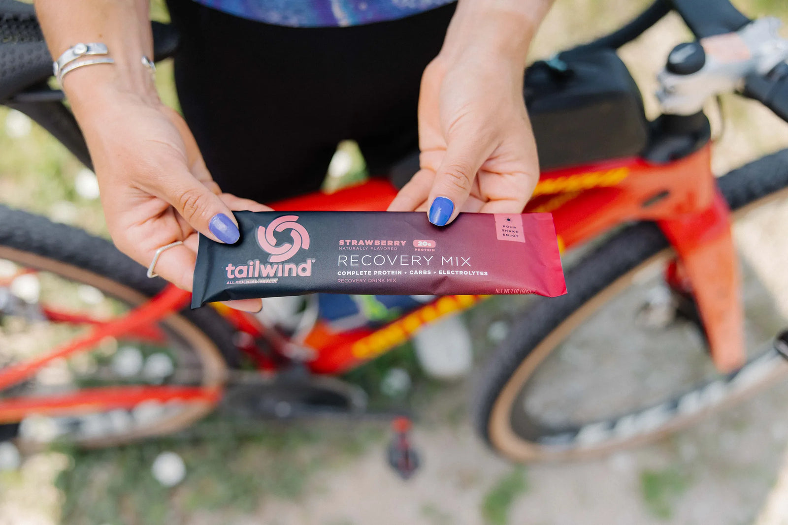 Person holding Tailwind Recovery Mix strawberry protein powder next to a red bicycle outdoors