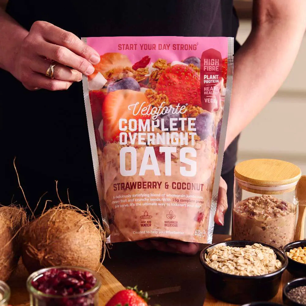 Veloforte Complete Overnight Oats Strawberry & Coconut pouch held above jars of oats and fruit